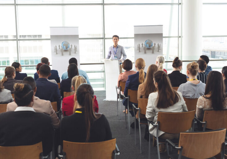 Front view of young Asian businessman speaking in front of business people sitting at business seminar in office building